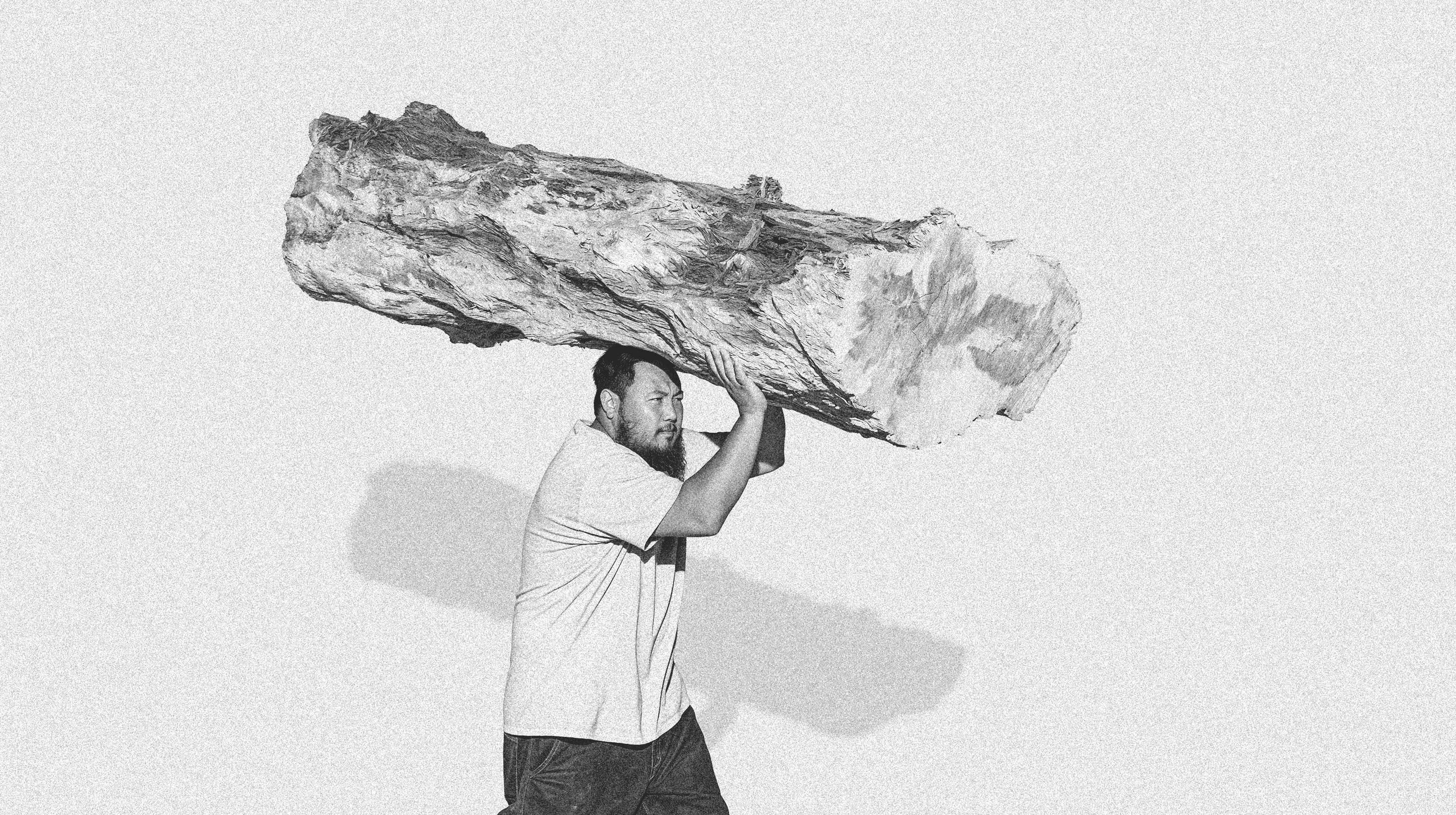 A man carrying a large rough log on his shoulder, black-and-white, against a soft grey wall