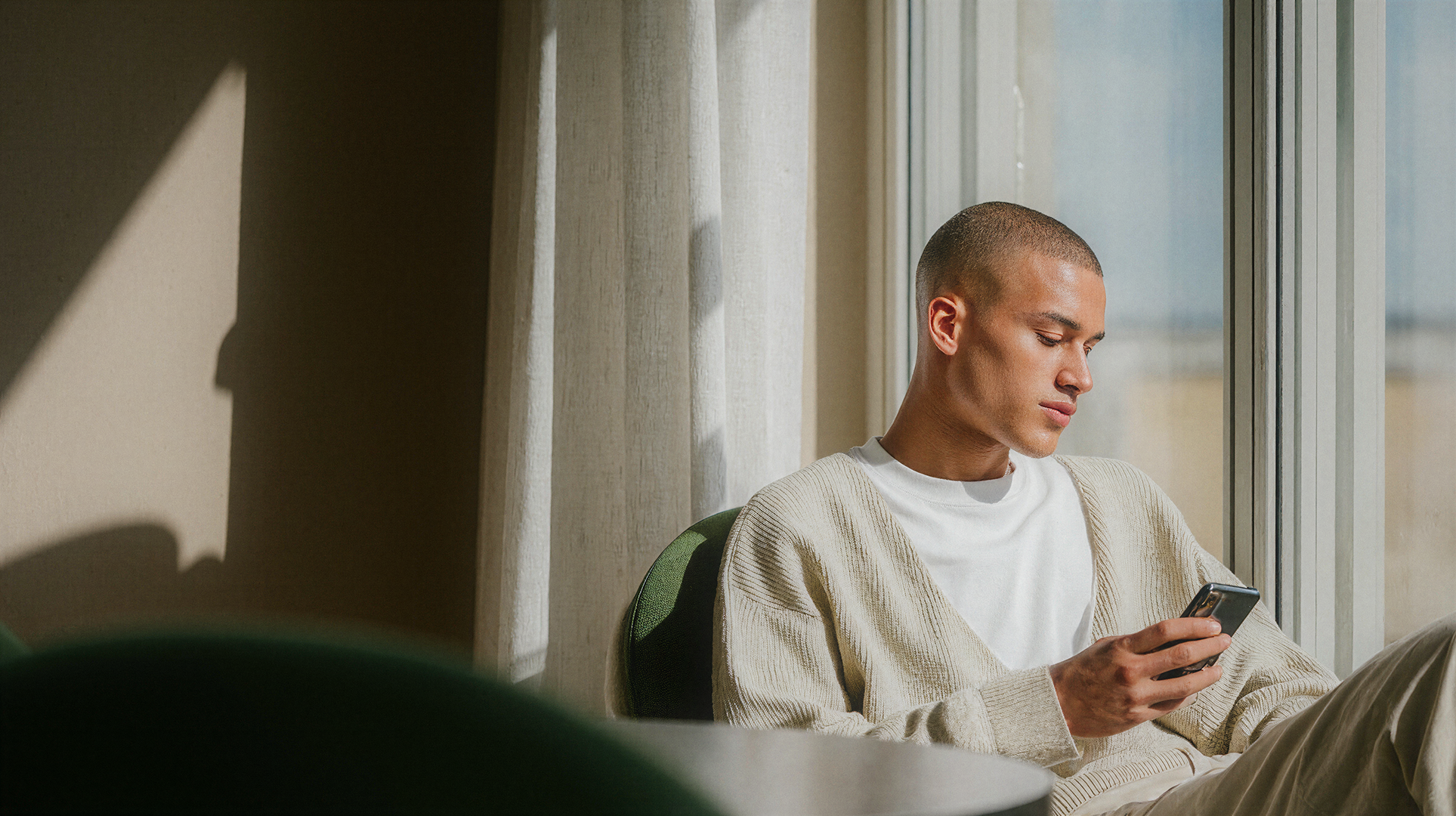 Young man seated by sunlit window using his phone
